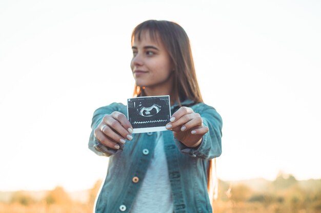 日没で超音波写真を保持し、彼女の腹を抱きしめる若い妊娠中の女性。妊娠4ヶ月。マタニティコンセプト。