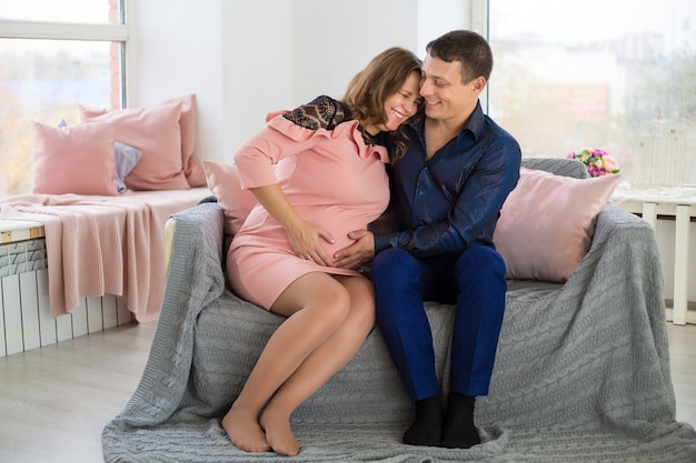 A young pregnant woman is sitting at a large window with her happy husband hugging her