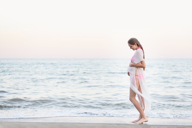 젊은 임신 한 여자는 해변에 서 있고 그녀의 배꼽을 포옹. 순간을 즐기고 있습니다.