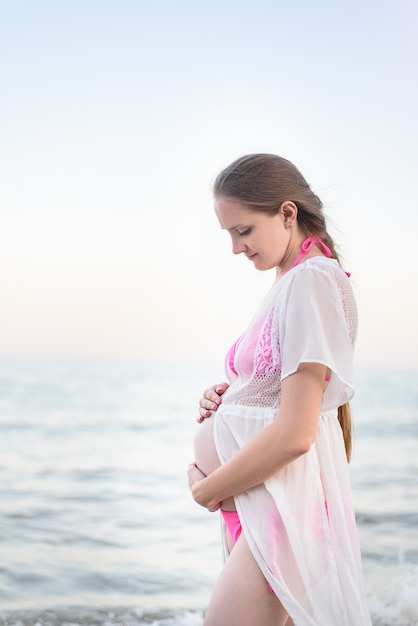 Young pregnant woman is standing on the seashore and hugging her belly