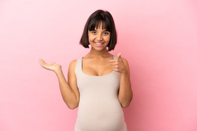 Young pregnant woman over isolated pink background holding copyspace imaginary on the palm to insert an ad and with thumbs up