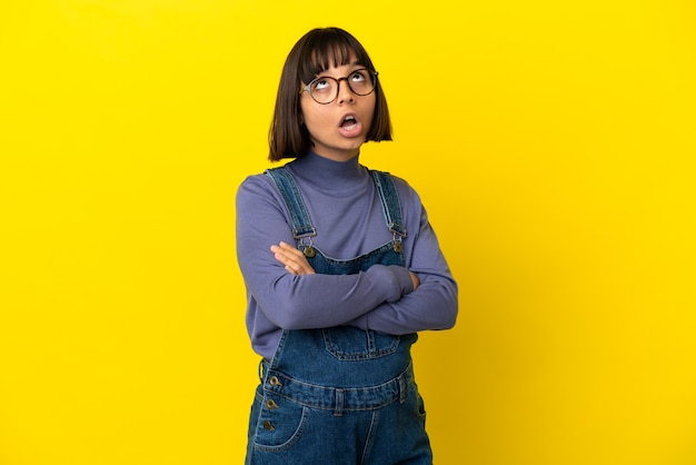 Young pregnant woman over isolated yellow background looking up and with surprised expression