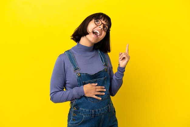 Young pregnant woman over isolated yellow background pointing up and surprised