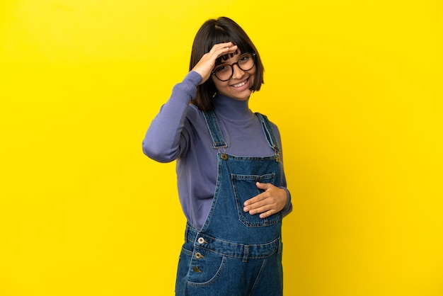 Young pregnant woman over isolated yellow background saluting with hand with happy expression