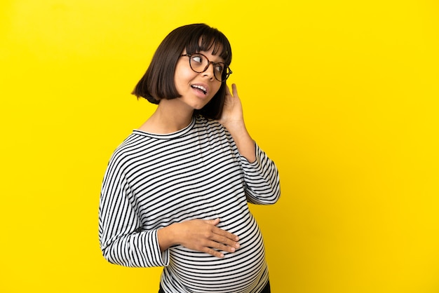 Young pregnant woman over isolated yellow wall listening to something by putting hand on the ear