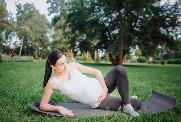 Young pregnant woman lying on yog mate and doing workout outside in park. She hold one leg in front another. One hand is on belly.