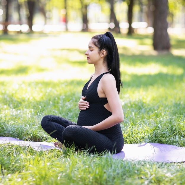 A young pregnant woman meditates sitting in a lotus position on a mat in the park