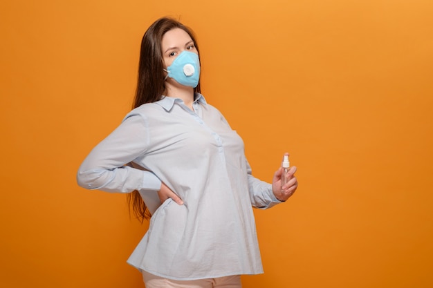 Young pregnant woman on orange wall in protective medical mask and spray antiseptic in hand