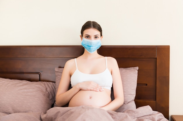 Young pregnant woman in protective medical mask is staying in bed because of coronavirus. Quarantine time. Health care concept