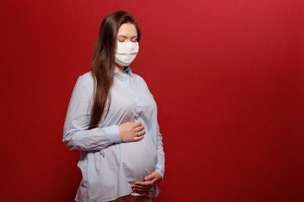 Young pregnant woman on red wall in protective medical mask holds on stomach