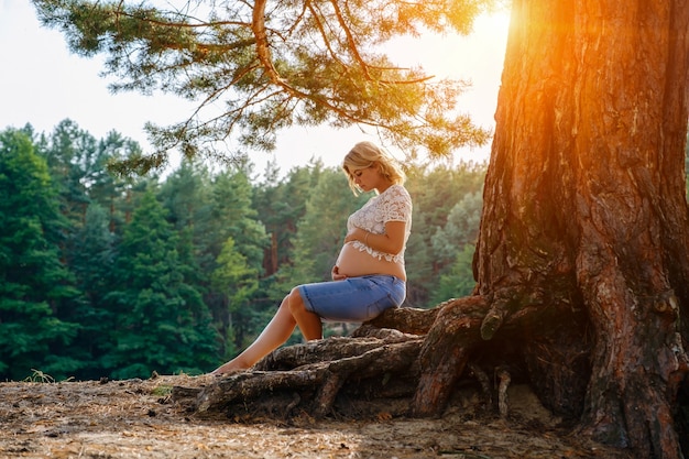 Young pregnant woman sits on the roots under a tree in the forest