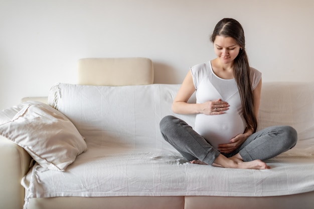 白いソファに座って、腹に手を保つ若い妊婦