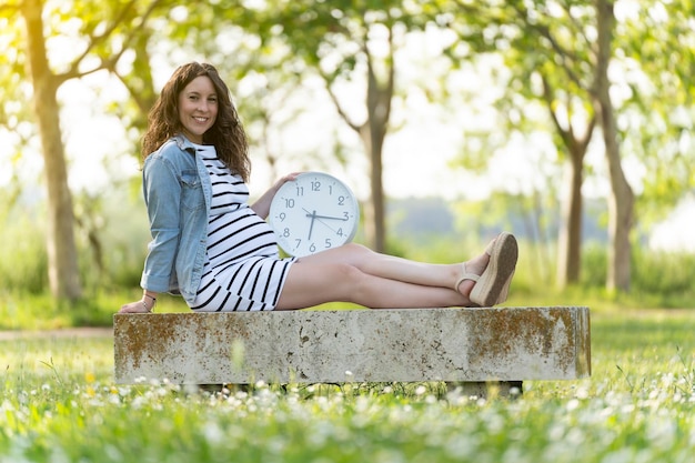 Young pregnant woman sitting with a clock in the park and looking to the camera