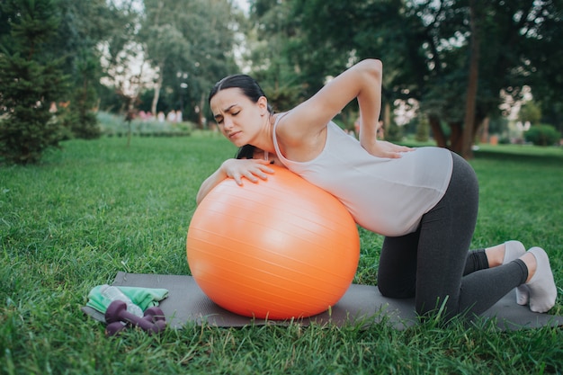 Young pregnant woman suffeer from pain in back. She stand in knees in park. Model lean to orange fitness ball.