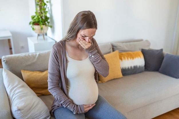 自宅で中毒に苦しんでいる若い妊婦。居間のソファに座っている間、手で口を覆うつわりを持っているマタニティ女性。お母さんは胎児を期待しています