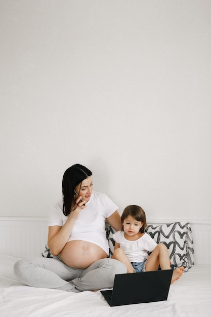 Young pregnant woman together with her little daughter sitting on the bed using gadgets