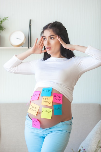 Young pregnant woman trying don`t forget stuff to hospital. Expecting female with notes on her stomach.