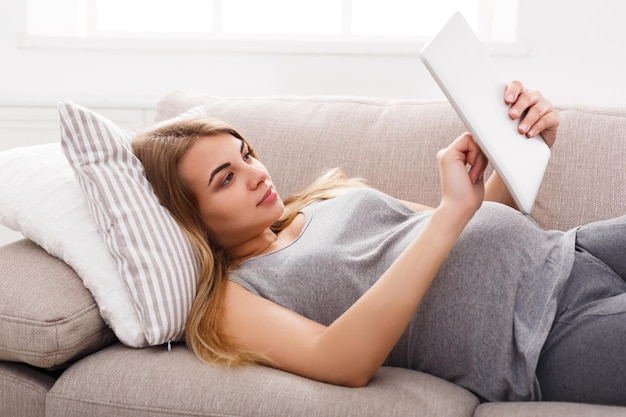 Young pregnant woman using digital tablet. Expectant blonde texting with tablet sitting on the floor. Networking, modern technologies, pregnancy, communication concept