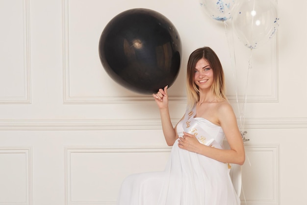 A young pregnant woman in a white dress hugs belly holding a black balloon isolated white neoclassical wall