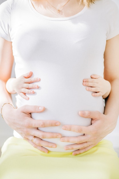 Young pregnant woman in white t-shirt and child hands touching her stomach