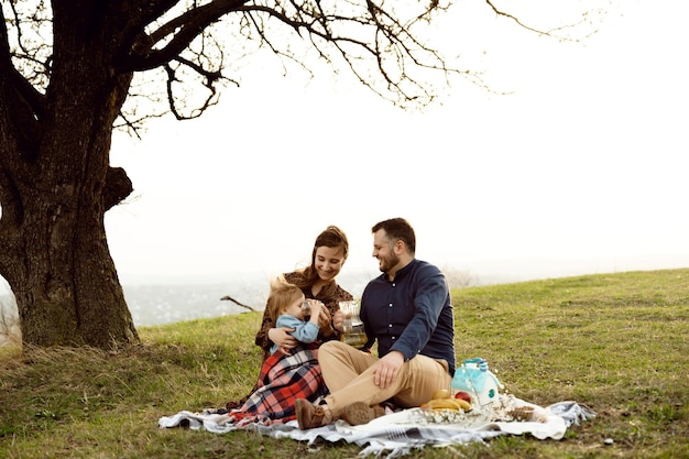 ハンサムな夫と小さな女の子を持つ若い妊婦は日没でピクニックにいます夫と妻は笑ってジュースを飲みます家族の時間