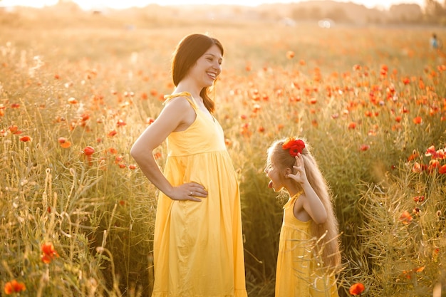 A young pregnant woman with her daughter in yellow dresses are standing in a poppy field Family relationships