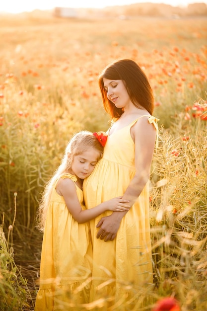 A young pregnant woman with her daughter in yellow dresses are standing in a poppy field Family relationships