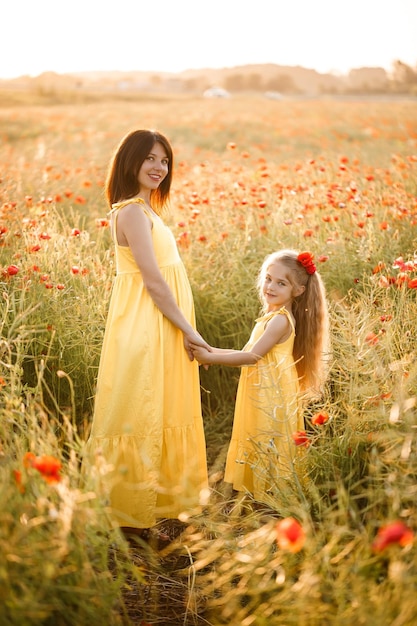 A young pregnant woman with her daughter in yellow dresses are standing in a poppy field Family relationships