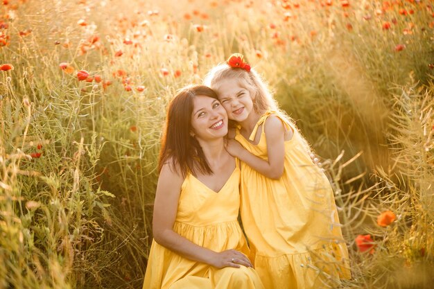 A young pregnant woman with her daughter in yellow dresses are standing in a poppy field Family relationships