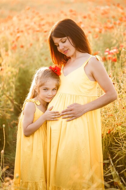 A young pregnant woman with her daughter in yellow dresses are standing in a poppy field Family relationships