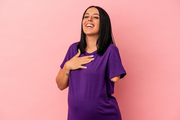 Young pregnant woman with one arm isolated on pink background laughs out loudly keeping hand on chest.