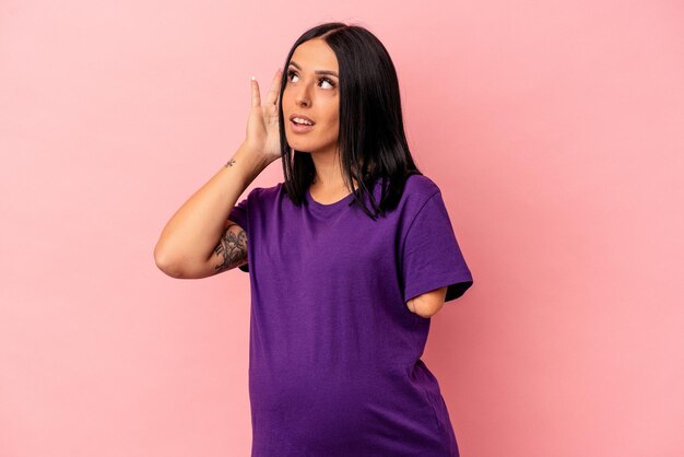 Young pregnant woman with one arm isolated on pink background trying to listening a gossip.