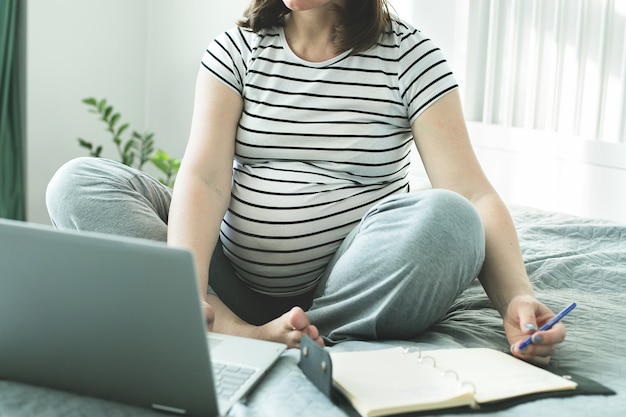 Young pregnant woman working from home office. Freelancer using laptop, notebook and the Internet. Workplace in bedroom. Concept of female business, career, home education, shopping online.Coronavirus work at home