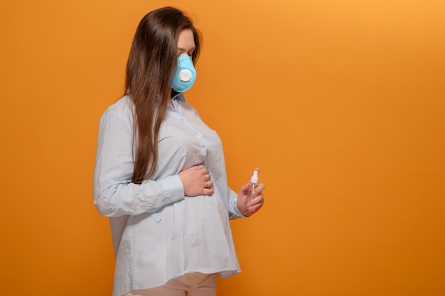 Giovane donna incinta sulla parete gialla in maschera medica protettiva e spray antisettico in mano