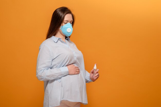 Young pregnant woman on yellow wall in protective medical mask and spray antiseptic in hand