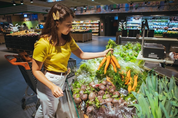 Молодая красивая взрослая женщина берет морковь с полки супермаркета