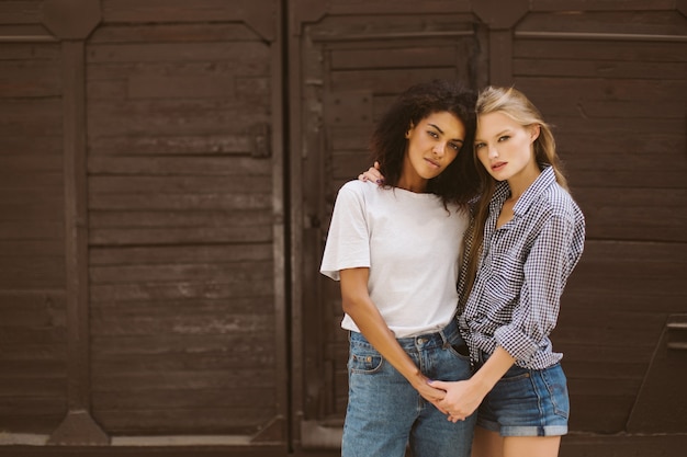 Young pretty african american woman with dark curly hair in T-shirt and jeans and attractive woman with blond hair in shirt and denim shorts dreamily  while spending time together