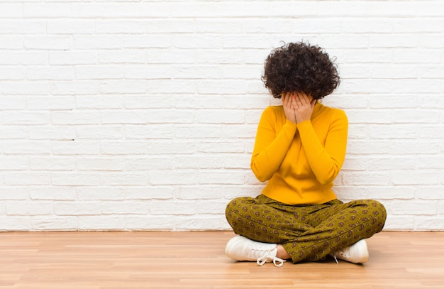 Young pretty afro woman covering eyes with hands with a sad, frustrated look of despair, crying, side view sitting on the floor