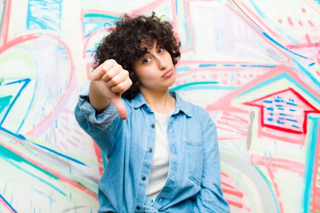 Young pretty afro woman feeling cross angry annoyed disappointed or displeased showing thumbs down with a serious look against graffiti wall