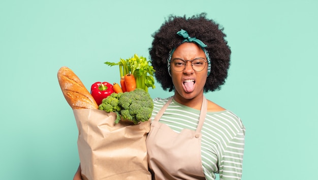 Giovane bella donna afro che si sente disgustata e irritata, sporge la lingua, non gli piace qualcosa di brutto e schifoso e tiene in mano un sacchetto di verdure