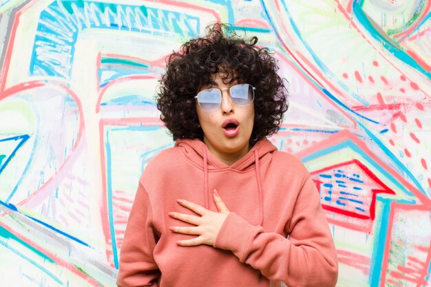 Photo young pretty afro woman feeling shocked and surprised, smiling, taking hand to heart, happy to be the one or showing gratitude against graffiti wall