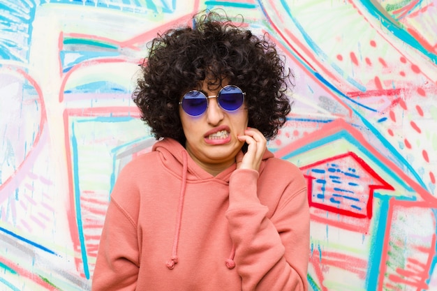 Photo young pretty afro woman feeling stressed, frustrated and tired, rubbing painful neck, with a worried, troubled look against graffiti wall