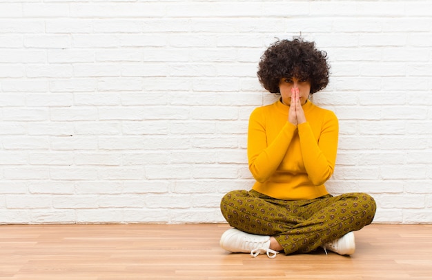 Young pretty afro woman feeling worried, hopeful and religious, praying faithfully with palms pressed, begging forgiveness sitting on the floor