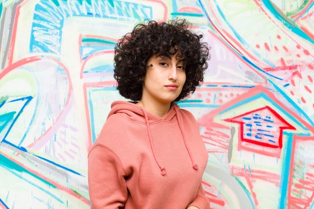 Young pretty afro woman looking proud, confident, cool, cheeky and arrogant, smiling, feeling successful against graffiti wall