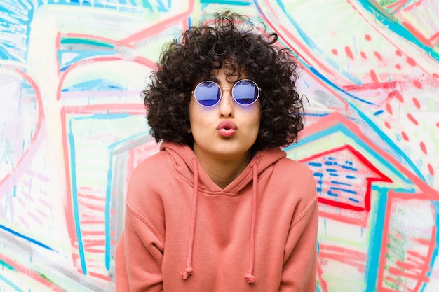 Young pretty afro woman pressing lips together with a cute, fun, happy, lovely expression, sending a kiss against graffiti wall