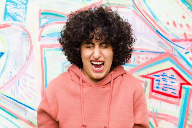 Photo young pretty afro woman shouting aggressively, looking very angry, frustrated, outraged or annoyed, screaming no against graffiti wall