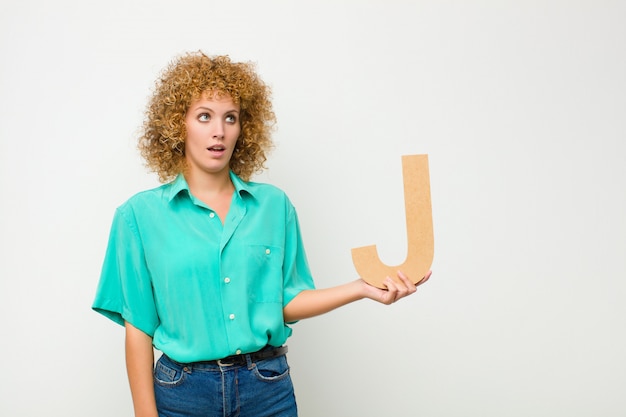 Foto giovane bella donna afro sorpresa, scioccata, stupita, con in mano la lettera j dell'alfabeto per formare una parola o una frase