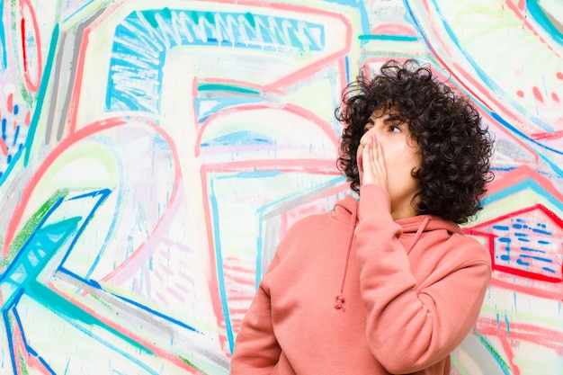 Young pretty afro woman yelling loudly and angrily to copy space on the side, with hand next to mouth against graffiti wall