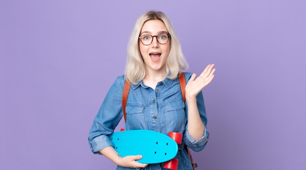 Young pretty albino woman feeling happy and astonished at something unbelievableand holding a skateboard