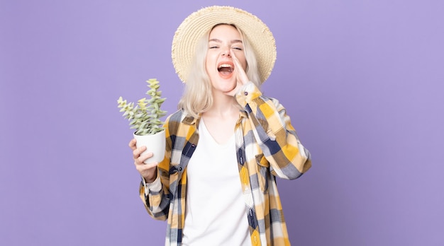Young pretty albino woman feeling happy,giving a big shout out with hands next to mouth and holding a houseplant cactus